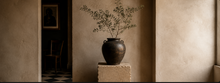 Dark ceramic vase with olive branches on a stone pedestal in a warm minimalist interior