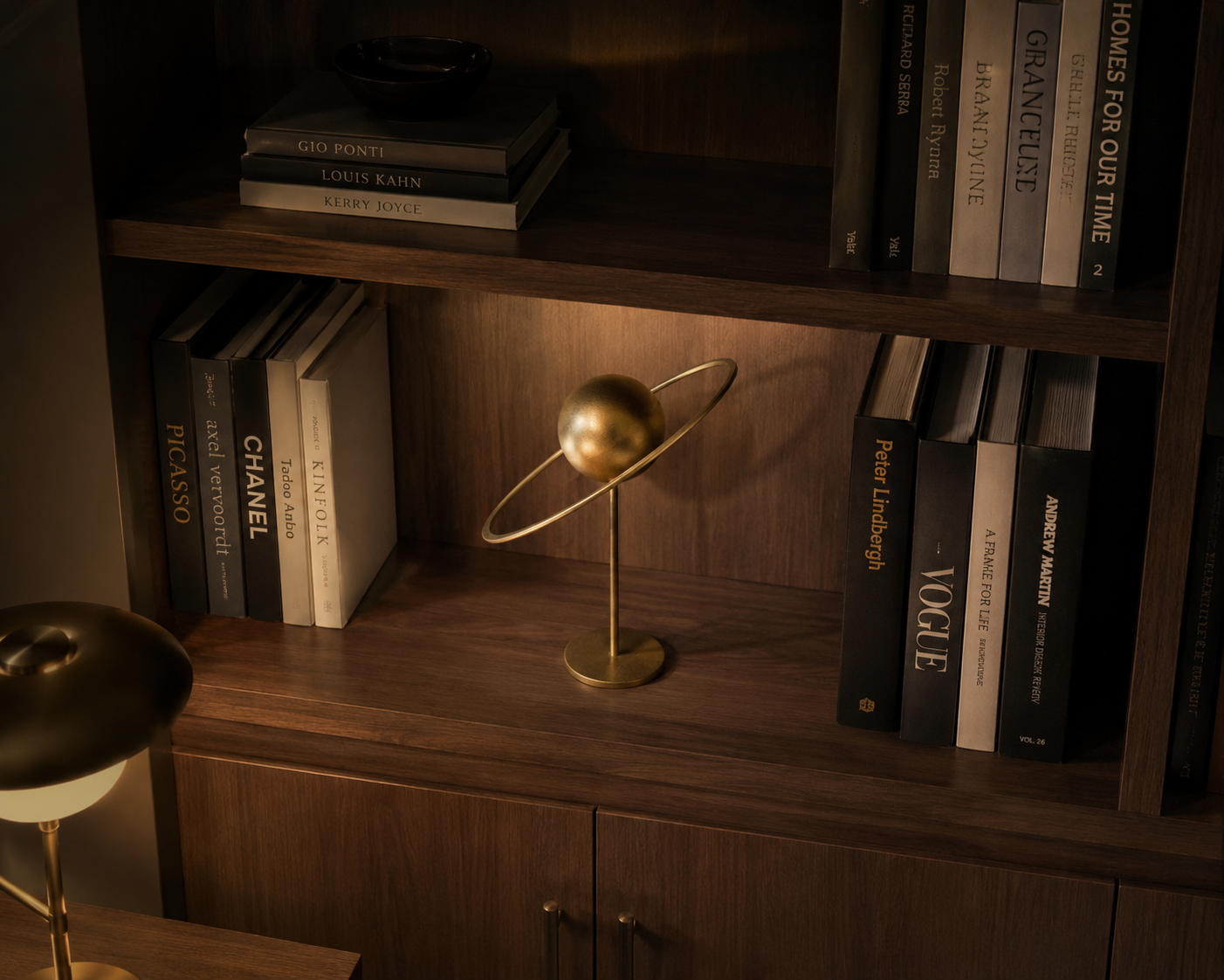 Gold iron planetary sculpture surrounded by design books on a warm wood library shelf at night.