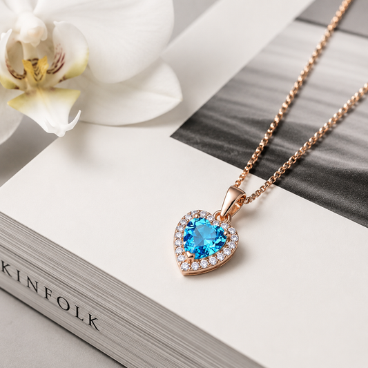 Rose gold cubic zirconia heart drop pendant with blue crystal, resting on a book beside an orchid