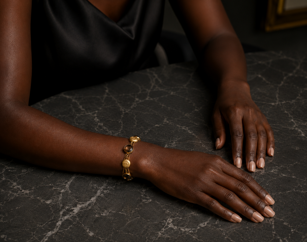 Black woman wearing an elegant gold bracelet with smoky stones on a dark marble table.